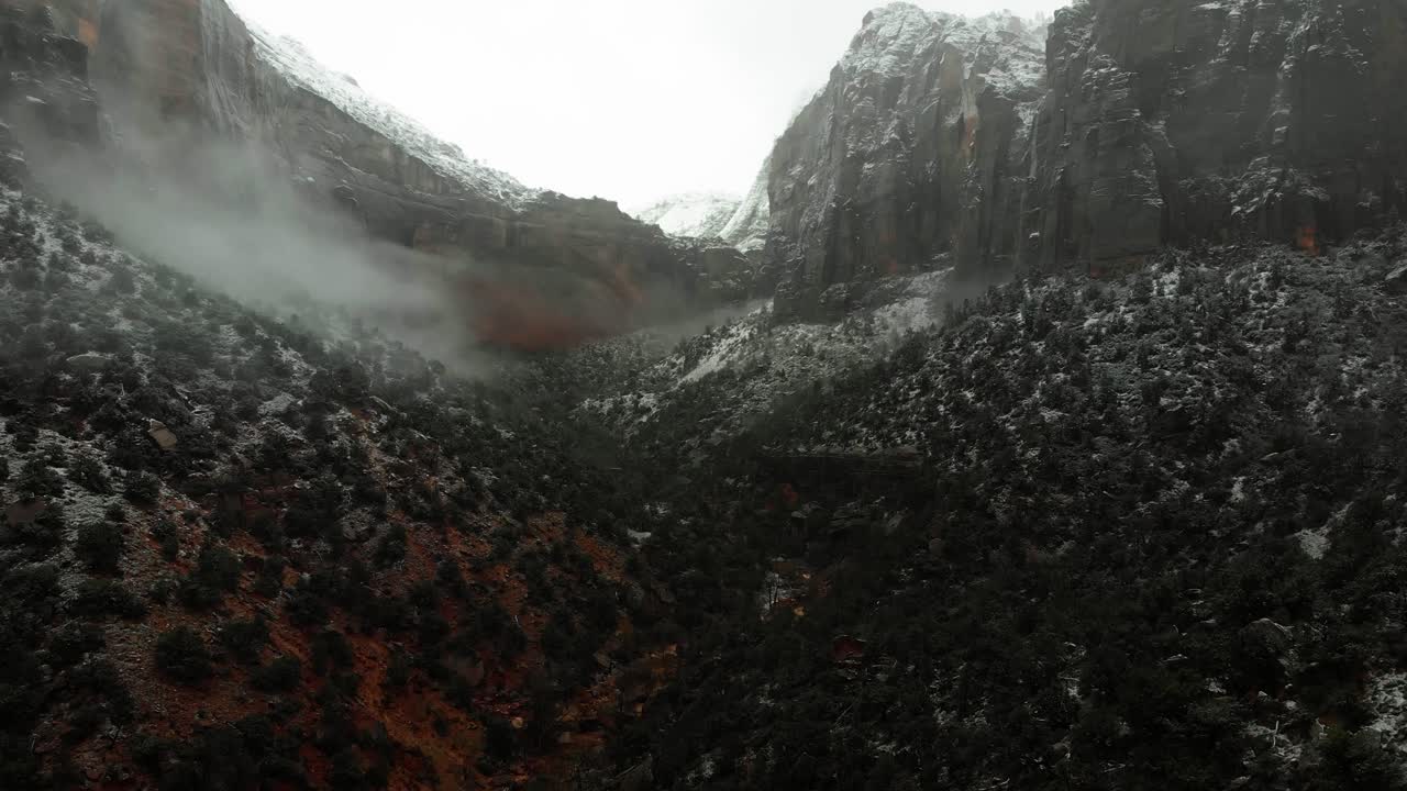 Drone shot descending into the valley between two mountains covered in snow and surrounded by thick fog and clouds.