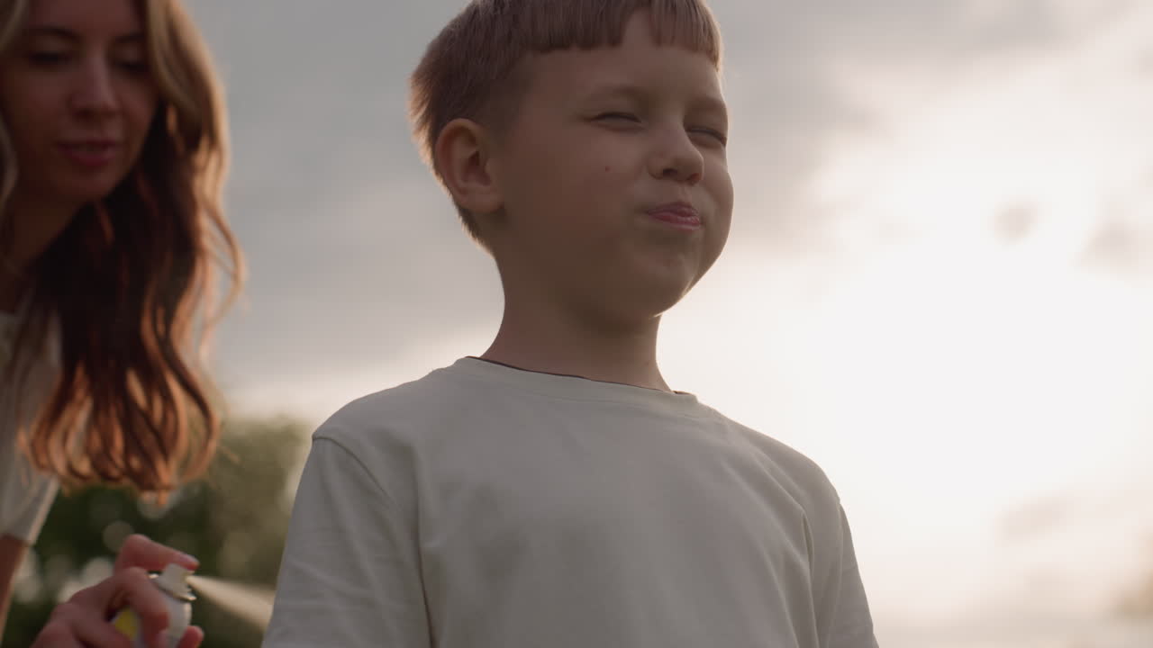 teenager tightening eyes and mouth while adult sprays back with cooling mist in bright sky outdoor moment, sun glow behind, playful discomfort and care captured during warm park outing