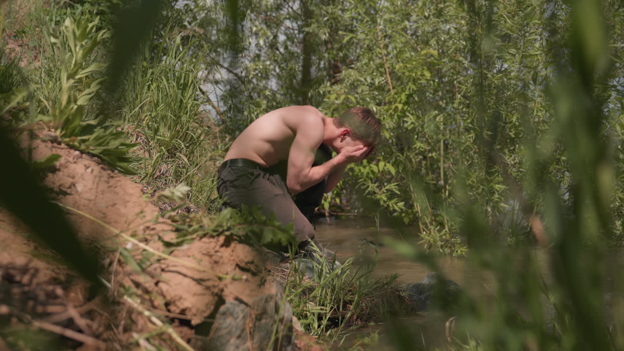 adventurer bending over riverbank to scoop water and wash muddy face, sunbeams filtering through surrounding trees, muscular torso and camo mud streaks visible as green foliage sways in breeze