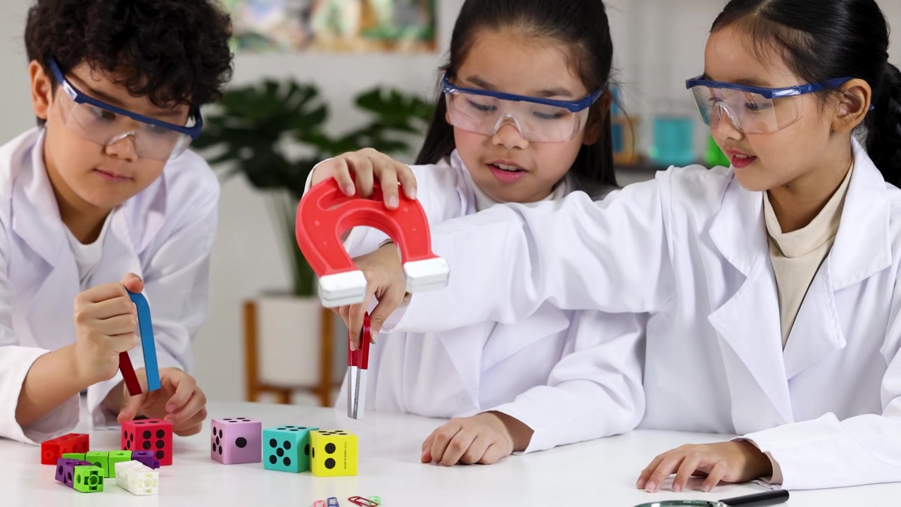 Anak-anak yang beragam mengenakan jas lab menggunakan magnet tapal kuda untuk menguji daya tarik pada balok-balok berwarna-warni.