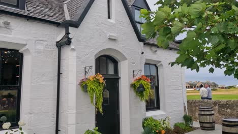 a quaint white cottage adorned with vibrant flowers and surrounded by lush greenery and rustic barrels.