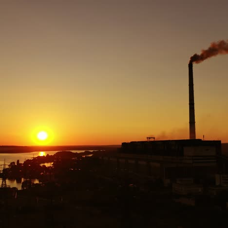 dark factory against the orange setting sun. factory pipes with smoke. dirty smoke pours out of chimneys into the air on the river background. aerial view.