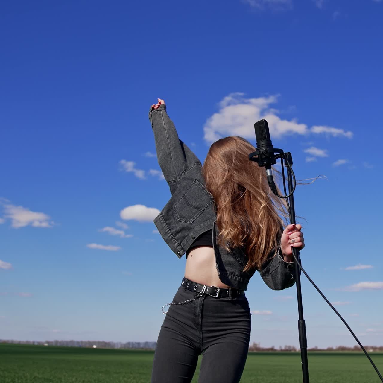 Young rock star dancing near microphone outdoors. Beautiful long-haired girl singing and moving her head with fluttering hair on blue sky background