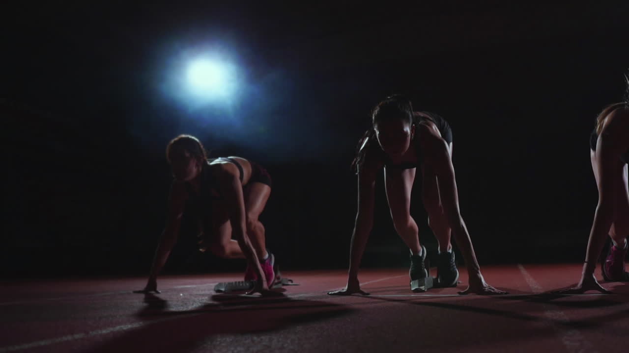 Three girls in black clothes are in the starting pads to start the race in the competition in the light of the lights and run towards the finish