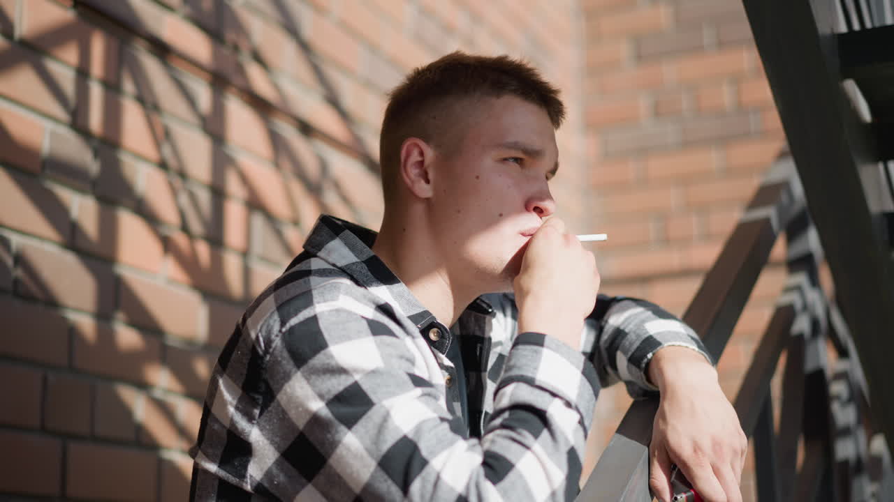 side view of student resting on iron railing smoking cigarette gazing thoughtfully into distant urban environment with check shirt warm sunlight casting shadows on brick wall and metal staircase