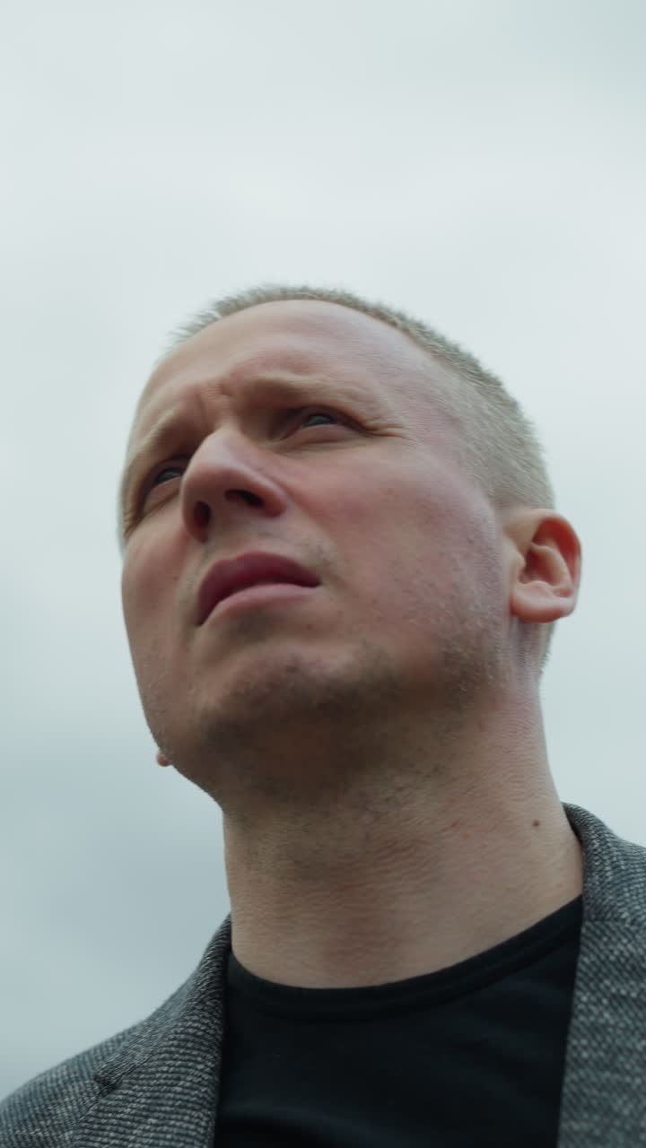 una vista de cerca de un hombre blanco envejecido en un traje gris y una camisa polo negra, mirando atentamente a la distancia con una expresión determinada, con un cielo nublado