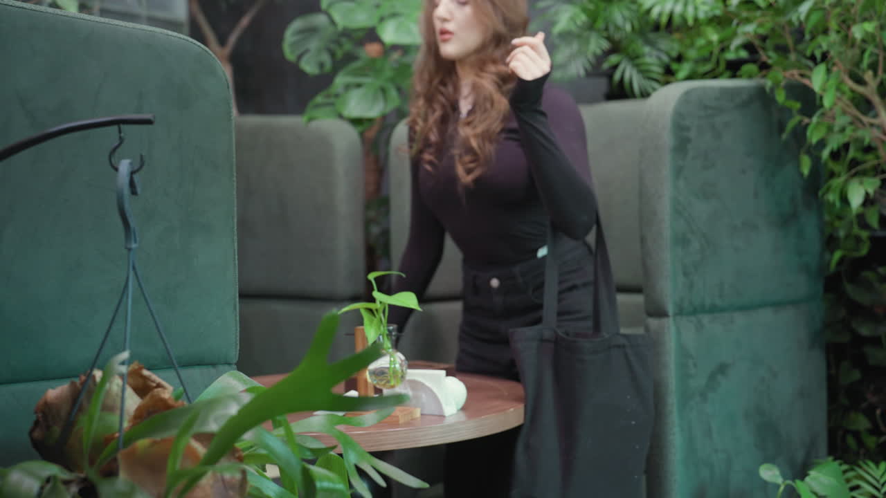 Close up of blonde woman in black outfit stepping into green booth area with hanging plant in foreground, soft indoor lighting and lush greenery create calm modern vibe in nature-inspired setting