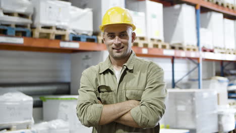 trabajador masculino sonriente parado en el almacén, cruzando los brazos y mirando la cámara