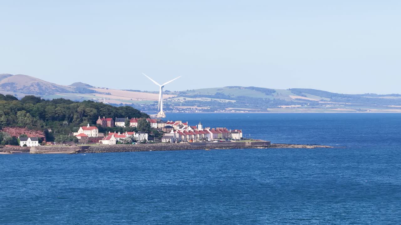 Wide shot of wind turbine spinning by seaside village, blue ocean, hills, bright daylight