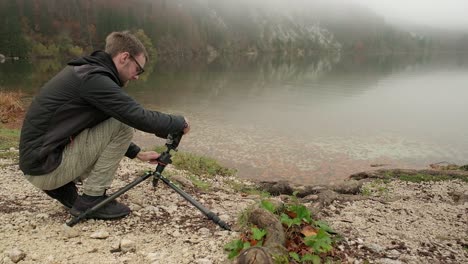 Fotograf,-Der-Seine-Kamera-Auf-Einem-Stativ-In-Bodennähe-Aufstellt,-Um-Neben-Einem-Seeufer-Zu-Fotografieren