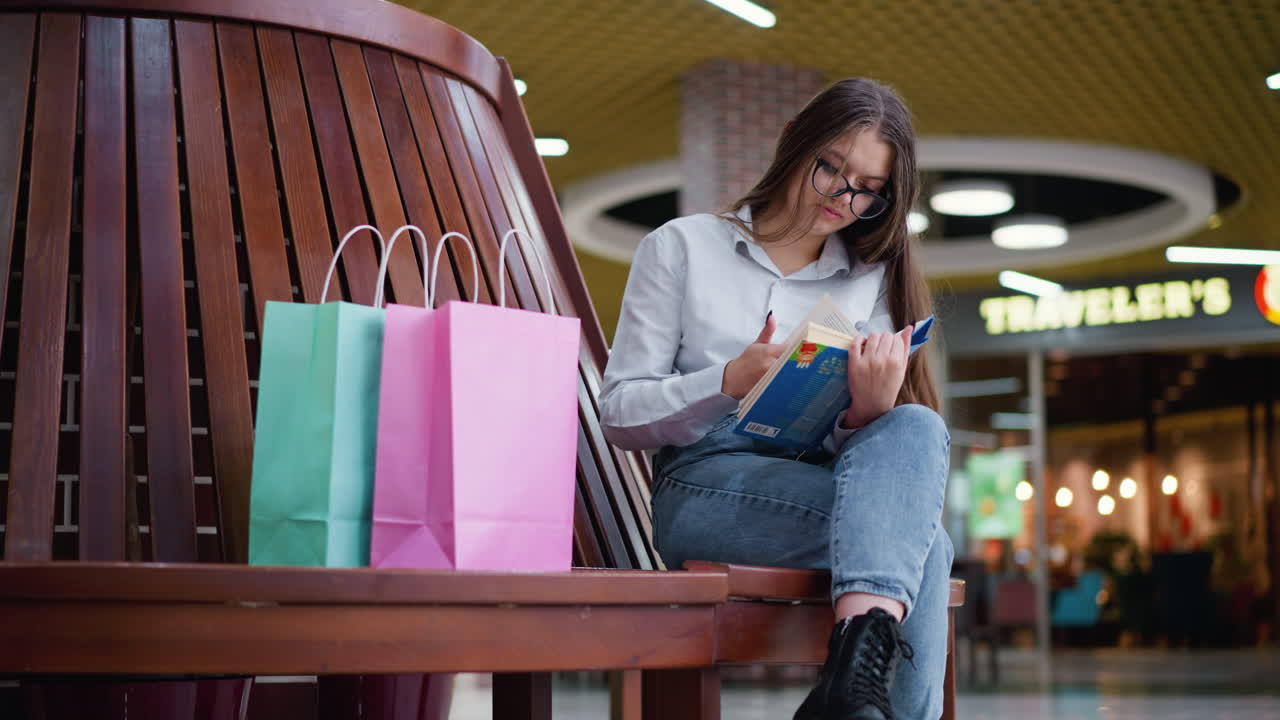 joven sentada en un banco de madera hojeando un libro, absorta en la lectura, con bolsas de compras a su lado en un vibrante entorno de centro comercial con luces bokeh y una acogedora cafetería en el fondo