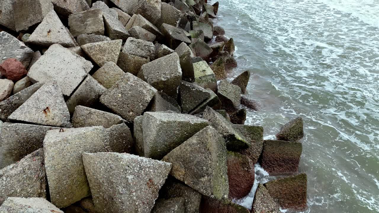 Aerial Fly Over jetty made of large, rectangular concrete blocks stacked on a sandy beach, with the ocean waves crashing in the background