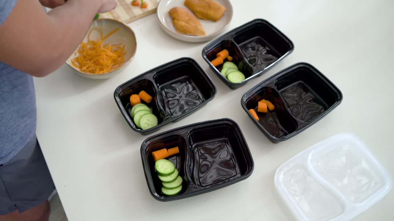 African American man prepping, placing carrots and cucumber into black containers with raw chicken
