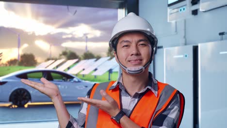 close up of asian male engineer with safety helmet smiling and pointing to side while standing with home energy storage system in a modern garage