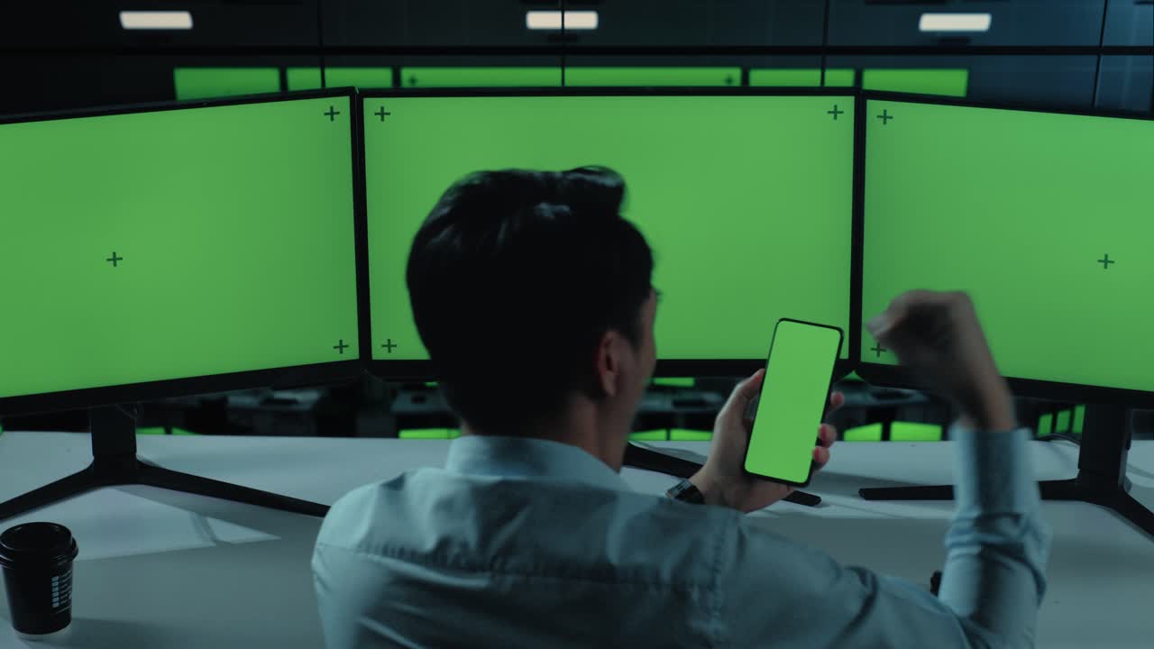 vista posterior de un hombre asiático feliz mirando la pantalla verde del teléfono móvil y celebrando mientras trabaja con múltiples monitores de computadora en la oficina