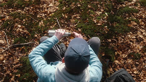 man-sitting-down-on-the-forest-floor,-pouring-coffee-in-his-cup-on-a-sunny-autumn-day