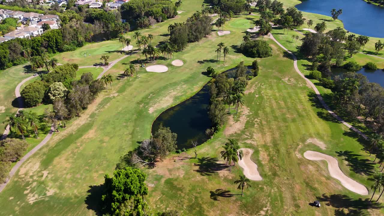 Sunny daytime drone flyover of lush golf fairway, sand bunkers, lakes, and surrounding neighborhood