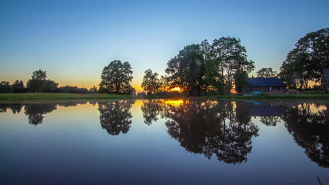 twilight to nighttime as the sunset fades and stars cross the sky reflecting off the surface of a lake by a countryside cottage - time lapse