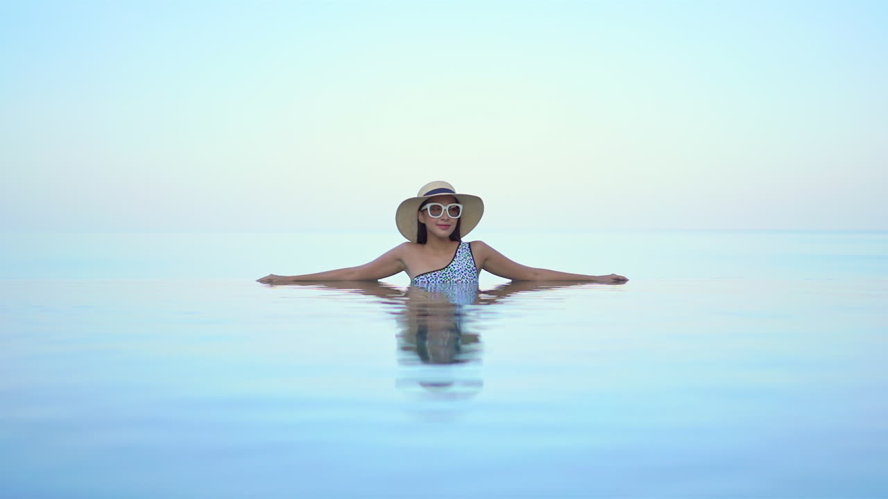 High-Key-Aufnahme einer schönen jungen lächelnden asiatischen Frau mit Hut und Sonnenbrille, die sich in einem Infinity-Pool entspannt.