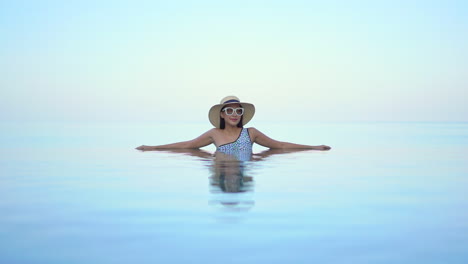 high key shot of beautiful young smiling asian woman with hat and sunglasses relaxing in infinity pool