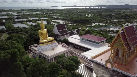 A-slow-orbital-shot-around-a-gold-statute-of-sitting-Buddha-and-a-temple-,-Thailand