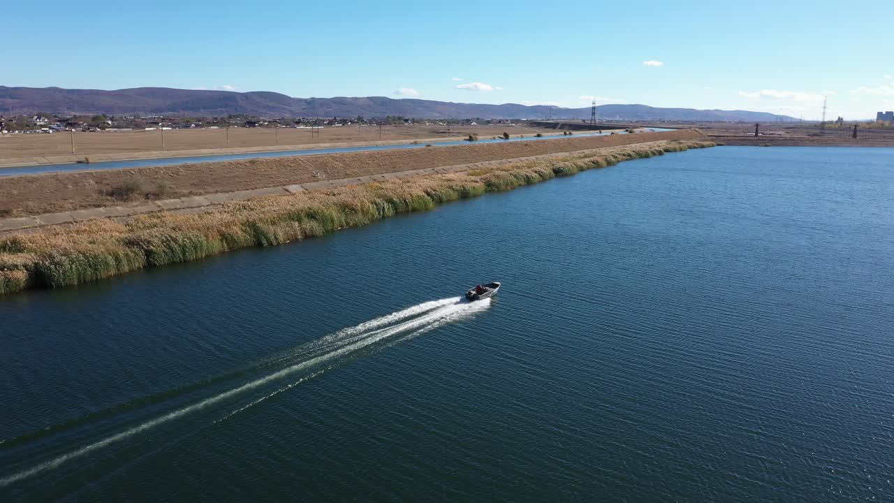 A drone tracks a speedboat racing across a lake on a bright, sunny day. The camera pans right, revealing an industrial power plant on the horizon behind the natural landscape. Bacau