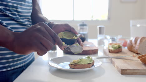 Hombre-Afroamericano-Poniendo-Aguacate-Sobre-Tostadas-En-Una-Cocina-Soleada,-Cámara-Lenta