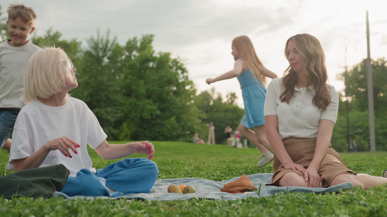 caretakers sitting on mat in grassy park laughing together while children run and play around them, summer afternoon, relaxed bonding, outdoor friendship, warm light creating joyful atmosphere