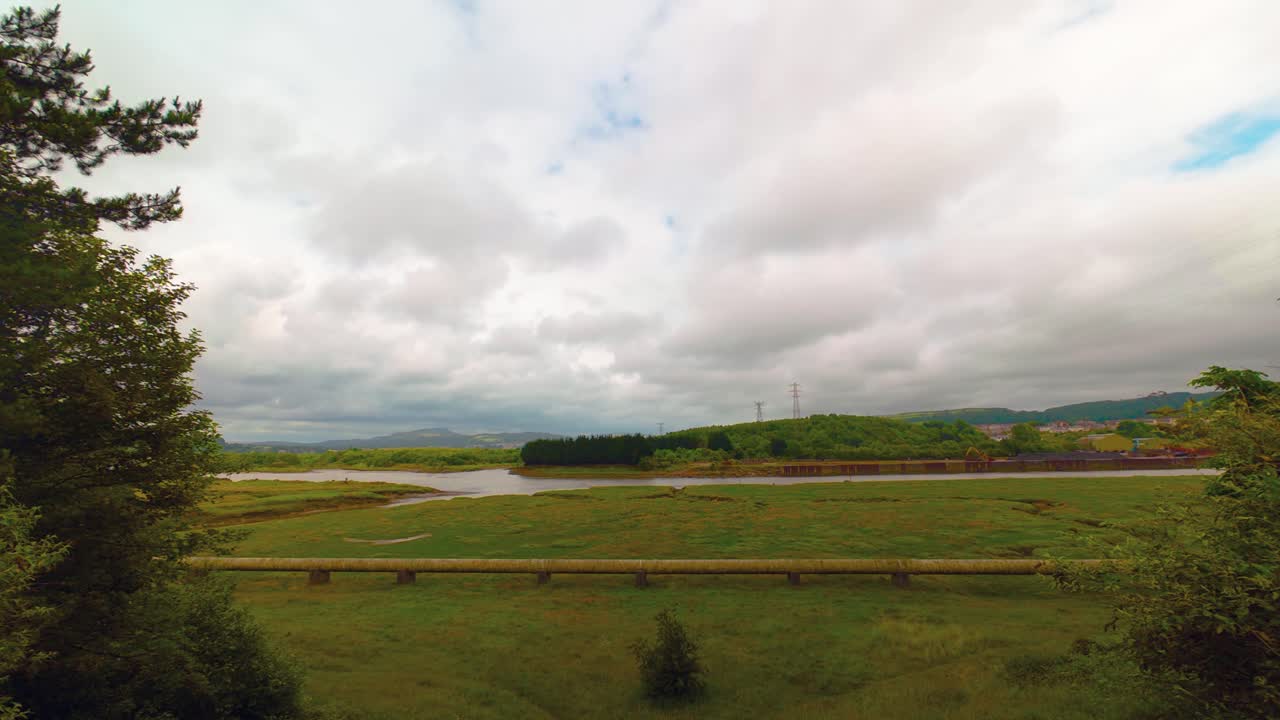 Slow Moving Timelapse of Wide Landscape with River Neath in Background with Gas Pipeline on Marshland Carrying Energy to Homes and Businesses with Thick Cloudscape.
