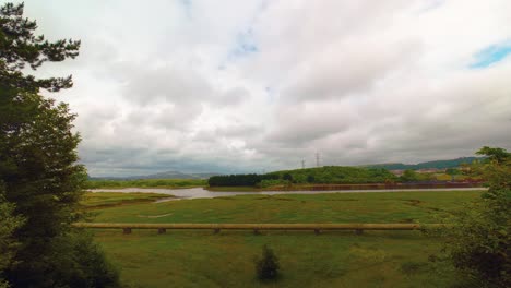 slow moving timelapse of wide landscape with river neath in background with gas pipeline on marshland carrying energy to homes and businesses with thick cloudscape.