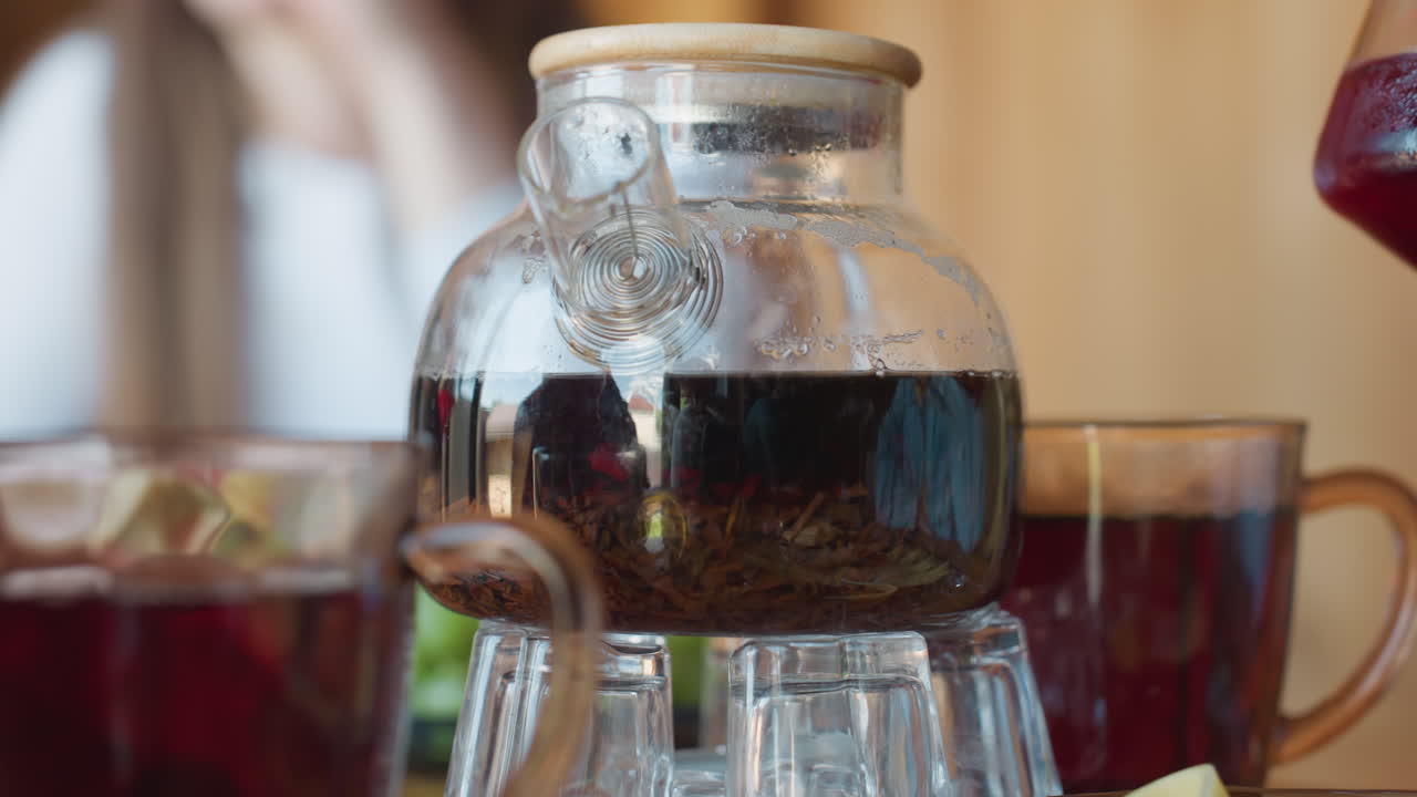 Detailed close up of dry herbs steeping inside transparent glass kettle with wooden lid, steam forming on surface, reflections of people and surroundings visible