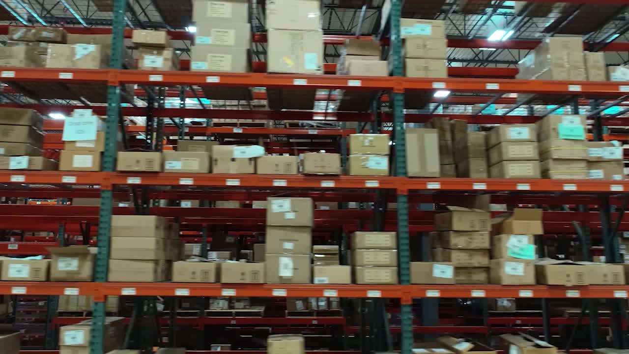 A lateral drone shot gliding left at a steady height, passing an underpass running through tall warehouse shelves stacked with boxes