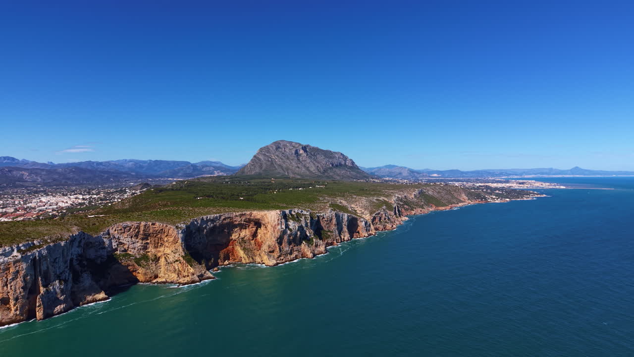 La penisola di Moraira sulla Costa Blanca, Spagna, che mostra drammatiche scogliere rocciose, il Mar Mediterraneo turchese e uno sfondo montuoso sotto un cielo azzurro e limpido.