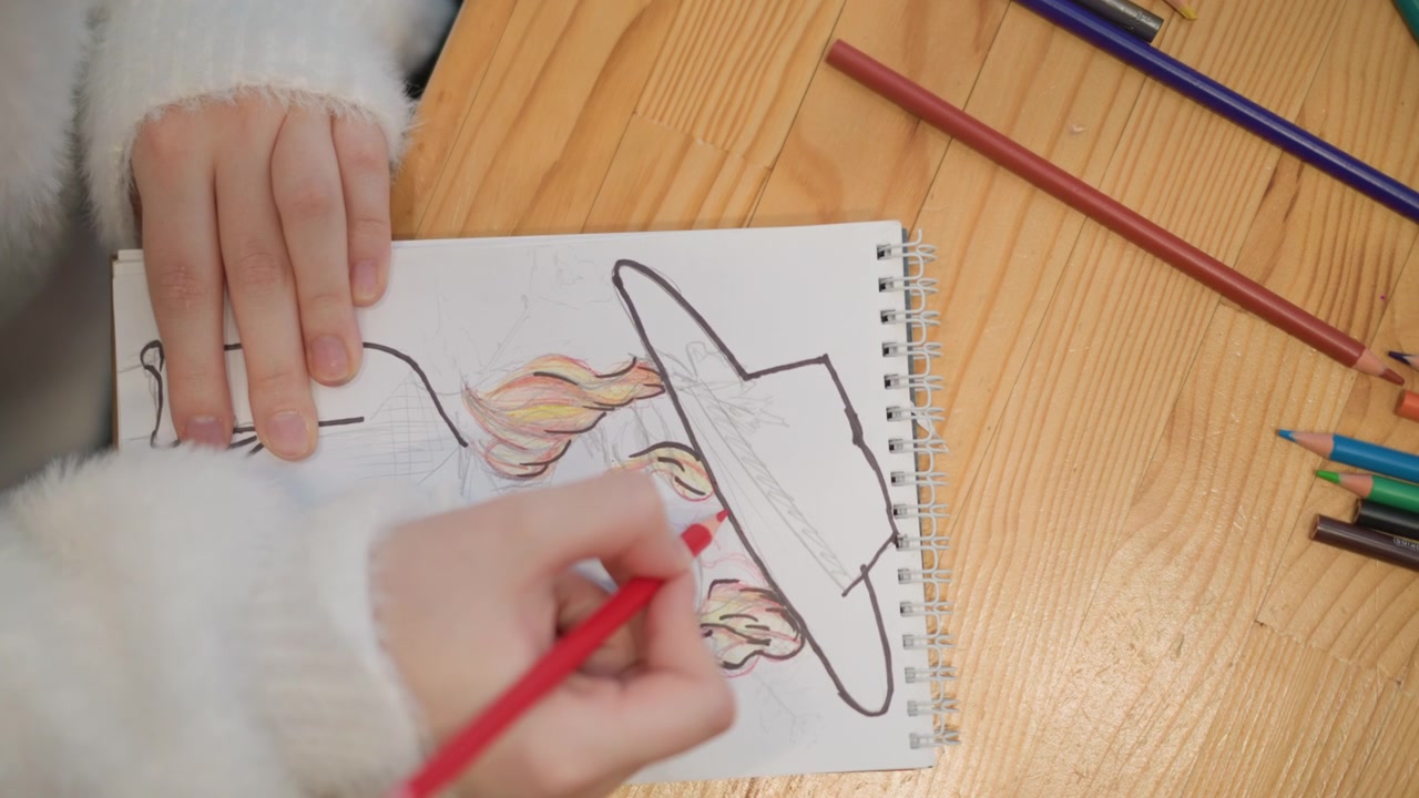 Vista ravvicinata dall'alto di un bambino che colora un volto illustrato con un cappello a tesa larga su un quaderno da disegno a spirale, la mano destra che riempie capelli e guancia con una matita rossa, mentre la mano sinistra tiene ferma la pagina su un tavolo di legno.
