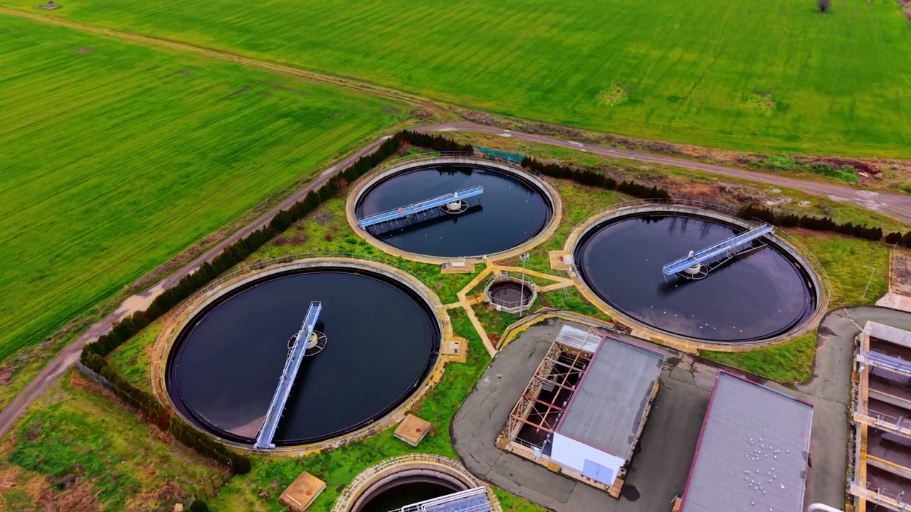 Aerial view of circular clarifier basins at wastewater treatment plant. Drone dark round sedimentation tanks, green lawn borders, blue pipes and white buildings.