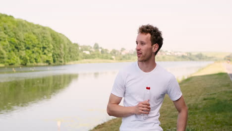 fitness young man resting and drinking water while leaning against metal railings in a park near the river after running workout