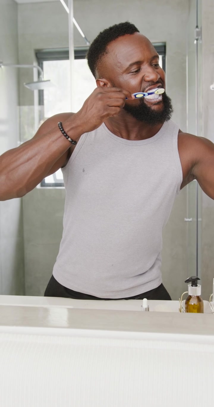 African American man in gray tank top brushing teeth with blue-white toothbrush at sink, rinsing
