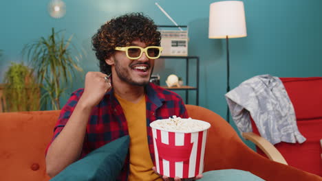 Indian-man-sits-on-couch-eating-popcorn-and-watching-interesting-TV-serial-sport-game-online-at-home