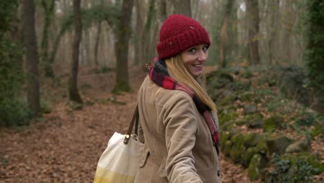 young woman walks in woods, turns and beckons to camera to follow her