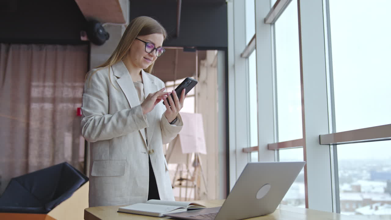 Good-looking lady standing in front of desk and taking notes on paper. Smiling woman makes a phonecall and looks at the screen.
