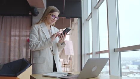 good-looking lady standing in front of desk and taking notes on paper. smiling woman makes a phonecall and looks at the screen.