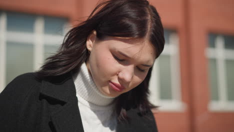 lady seated in black coat and whit turtle neck, focused on something, illuminated by sunlight, hair gently swaying in wind, background featuring red brick building