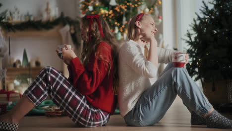 cheerful sisters singing and enjoying coffee while sitting on floor during christmas