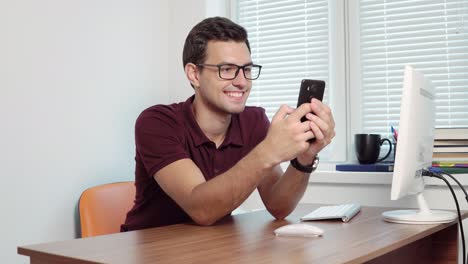 Man-looking-at-the-screen-of-his-phone-and-smiling,-texting-in-the-office-sitting-table.-Handsome-young-businessman-using