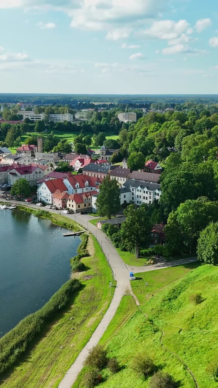 Aerial panorama capturing Talsi's historic center, featuring red tiled roofs, tranquil waterfront, verdant surroundings, and meandering path under summer sunlight