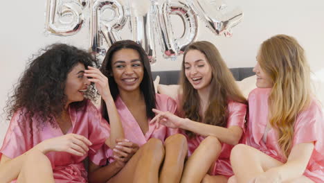 Grupo-De-Amigas-Multiétnicas-Vestidas-Con-Camisones-De-Seda-Rosa-Hablando-Y-Sentadas-En-La-Cama-Decoradas-Con-Globos-De-La-Palabra-&#39;novia&#39;-En-Una-Reunión-Nupcial-1