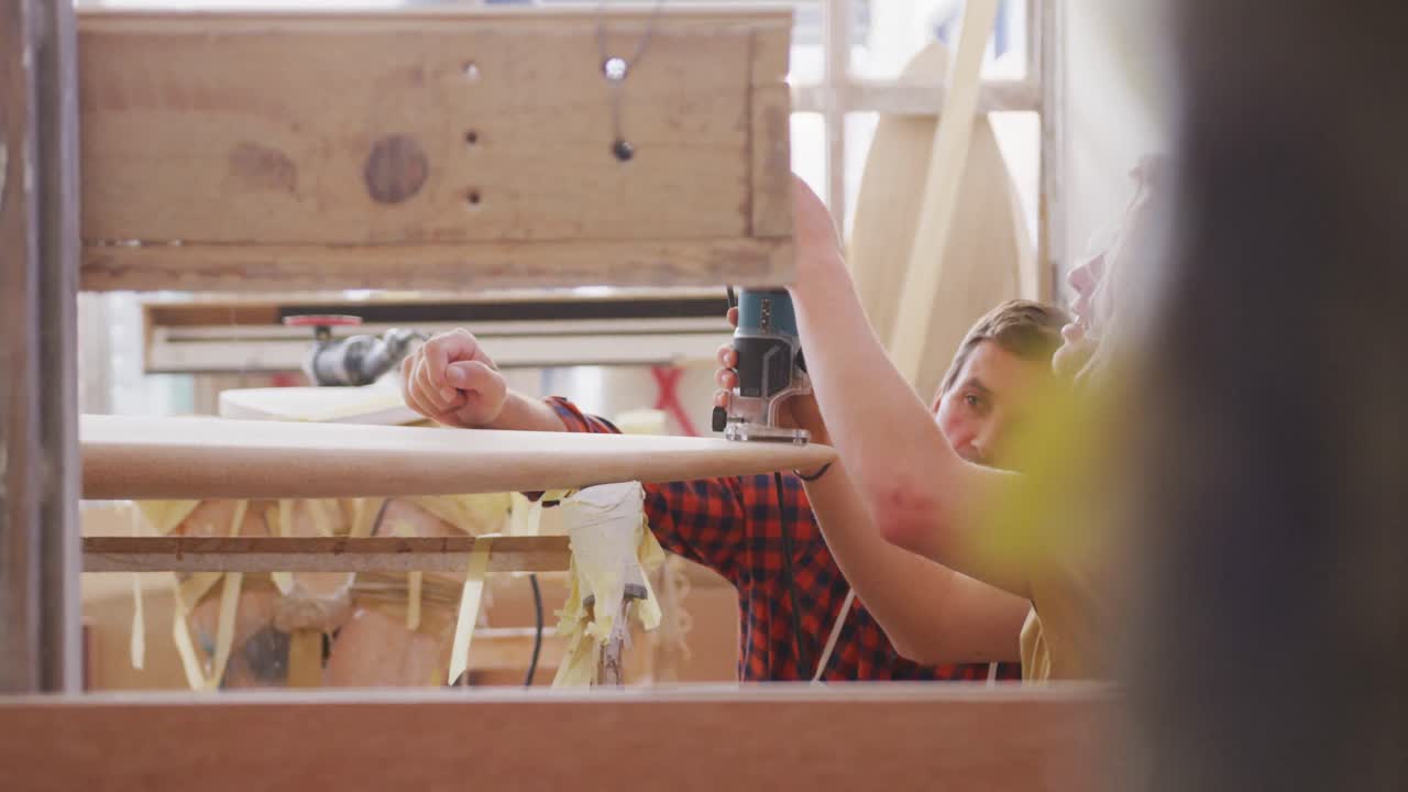 Two Caucasian male surfboard makers working in their studio and making a wooden surfboard together