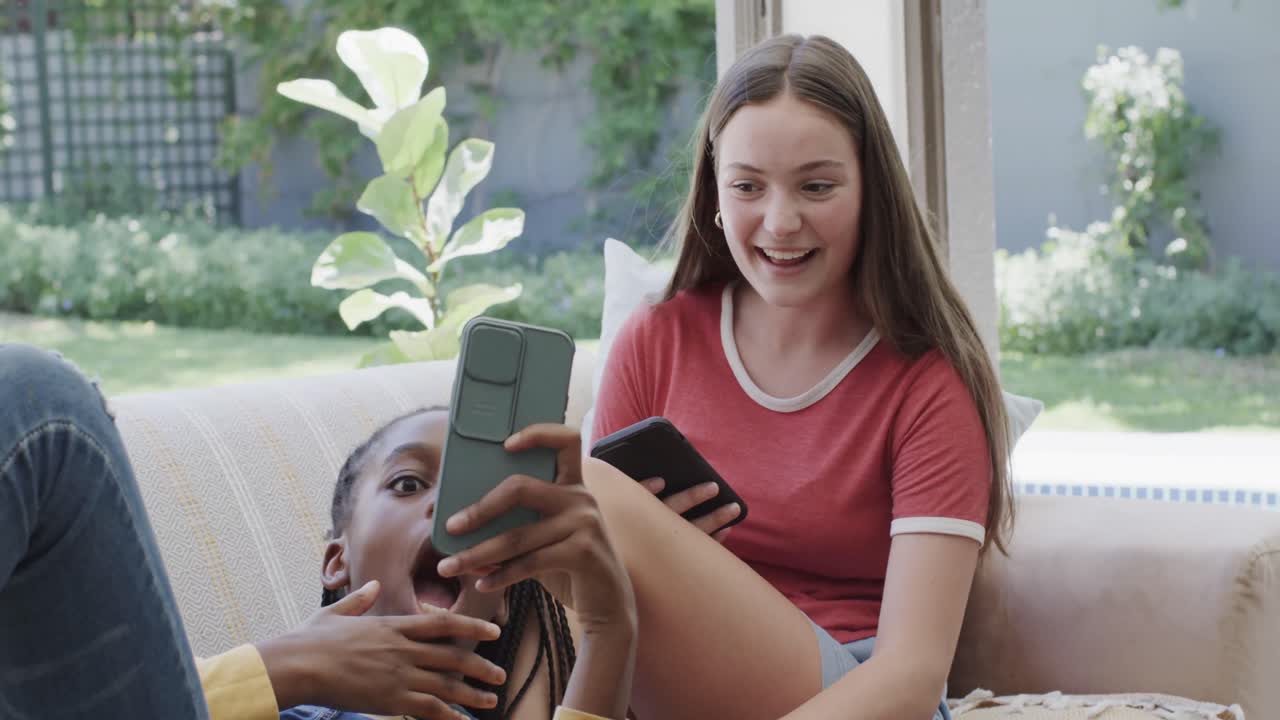 felices amigas adolescentes diversas acostadas en el sofá y usando teléfonos inteligentes en cámara lenta