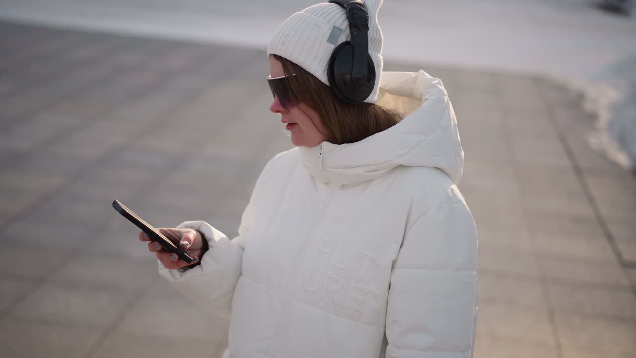 Student grooving and tapping smartphone screen while walking on tiled sidewalk in winter outfit with beanie headset and sunglasses under golden hour light in urban park setting scene
