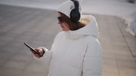 student grooving and tapping smartphone screen while walking on tiled sidewalk in winter outfit with beanie headset and sunglasses under golden hour light in urban park setting scene